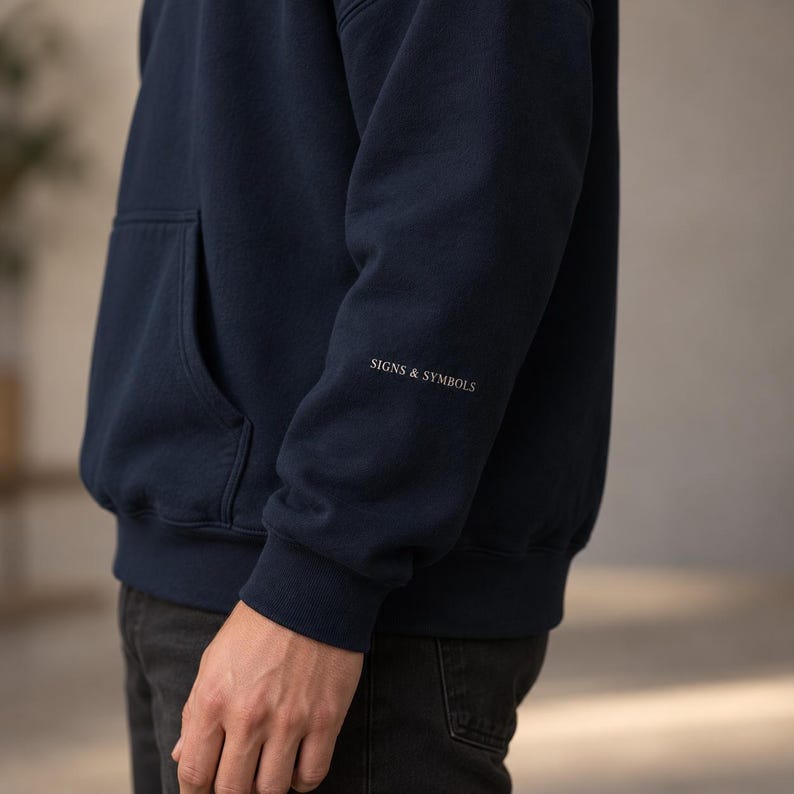 May include: A navy blue hooded sweatshirt featuring a front kangaroo pocket and the text SIGNS & SYMBOLS printed in a small white serif font on the lower portion of the left sleeve, finished with ribbed cuffs and a ribbed hem.