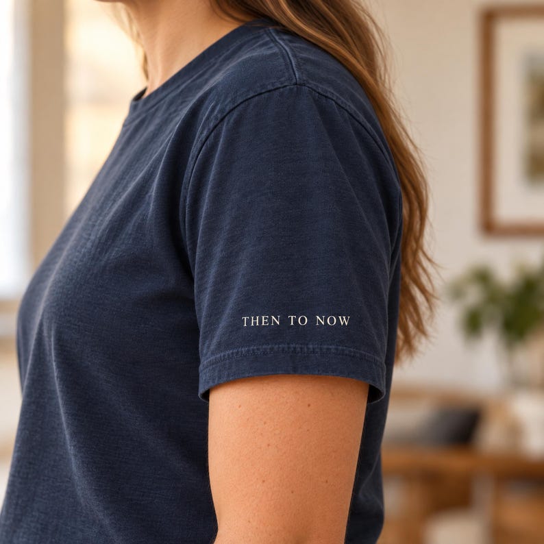 May include: A dark navy blue short-sleeved t-shirt with a crew neckline. The sleeve features the text THEN TO NOW printed in a small, white, sans-serif font. The fabric has a slightly textured, washed appearance throughout the garment.