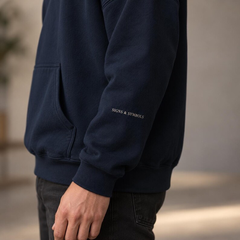 May include: A navy blue long-sleeve hooded sweatshirt featuring a front kangaroo pocket and the text SIGNS & SYMBOLS printed in a small, white, serif font on the lower portion of the left sleeve, just above the ribbed cuff.