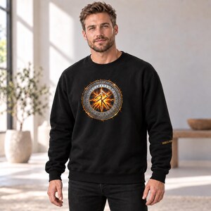 May include: A black long-sleeve sweatshirt featuring a circular graphic on the chest. The design displays a glowing orange rune symbol surrounded by a ring of Norse runes and a braided border. The text SIGNS & SYMBOLS is printed on the sleeve.