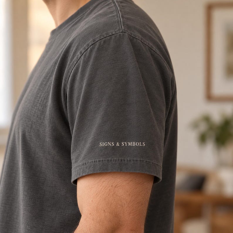 Pode incluir: Uma t-shirt de manga curta cinzento-carv&atilde;o com o texto SIGNS & SYMBOLS impresso numa fonte pequena, branca e sem serifas na parte inferior da manga esquerda. A pe&ccedil;a tem um decote redondo e uma costura vis&iacute;vel ao longo do ombro.