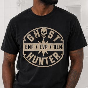 May include: A black short-sleeved t-shirt featuring a circular beige graphic with a skull icon at the top and the text GHOST HUNTER and EMF / EVP / REM printed in the centre. The design has a distressed, weathered appearance on the fabric.