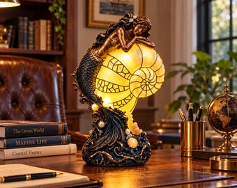 May include: A decorative table lamp featuring a bronze-toned mermaid resting on a large, glowing, translucent nautilus shell. The base is sculpted with dark blue, wave-like textures and small white pearl accents, emitting a warm, soft light.