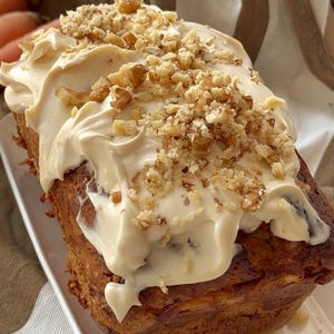 Puede incluir: Un pan rectangular de pastel de zanahoria cubierto con una capa gruesa de glaseado de queso crema de color beige claro y una generosa cantidad de nueces picadas, servido en un plato rectangular blanco con un paño marrón y blanco.