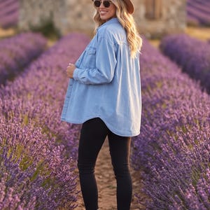 Op de afbeelding: Een lichtblauw spijkerhemd met lange mouwen en borstzakken, gecombineerd met een zwarte skinny jeans, beige veterschoenen, een lichtbruine fedora-hoed en een donkere ronde zonnebril in een veld met paarse lavendelbloemen.