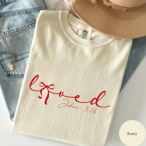 Op de afbeelding: Een ivoorkleurig T-shirt met korte mouwen en de rode cursieve tekst "loved", waarbij een strik in de letter "v" is verwerkt, met daaronder de tekst "John 3:16". Het label van het merk Comfort Colors is zichtbaar in de halslijn.