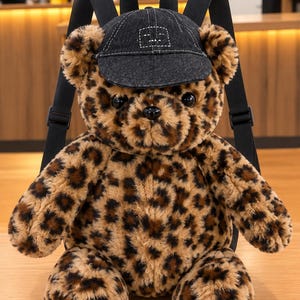 May include: A plush teddy bear backpack featuring a brown and black leopard print pattern. The bear wears a dark charcoal denim baseball cap with white stitching. It has black plastic eyes and nose, with black adjustable shoulder straps attached.
