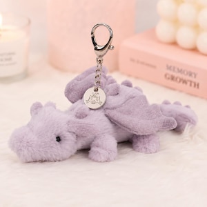 Pode incluir: Um porta-chaves de dragão de peluche cor de lavanda com pelo macio, asas pequenas e um fecho mosquetão de metal prateado. Um pendente circular prateado está preso à corrente, exibindo um desenho gravado de um gato com um chapéu de festa.