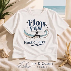 Puede incluir: Una camiseta blanca de manga corta con un gráfico de una persona en postura de yoga. El texto Flow First, Hustle Later aparece en azul oscuro. El diseño incluye una onda multicolor, pequeñas estrellas y el texto Ink & Ocean Studio.