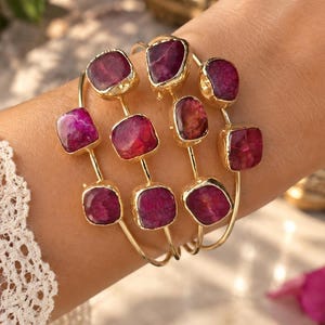 Op de afbeelding: Een set van drie goudkleurige draadarmbanden, elk voorzien van meerdere onregelmatige, gefacetteerde magentakleurige stenen in gouden zettingen. De armbanden worden samen gedragen en tonen een gelaagd ontwerp met gepolijste metalen banden en roze edelstenen.