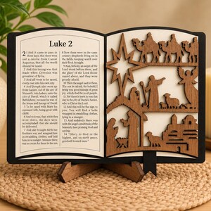 May include: A decorative wooden book display featuring the text of Luke 2 on the left page and a layered, laser-cut nativity scene on the right. The scene includes a star, camels, an angel, shepherds, sheep, and the holy family in a manger.