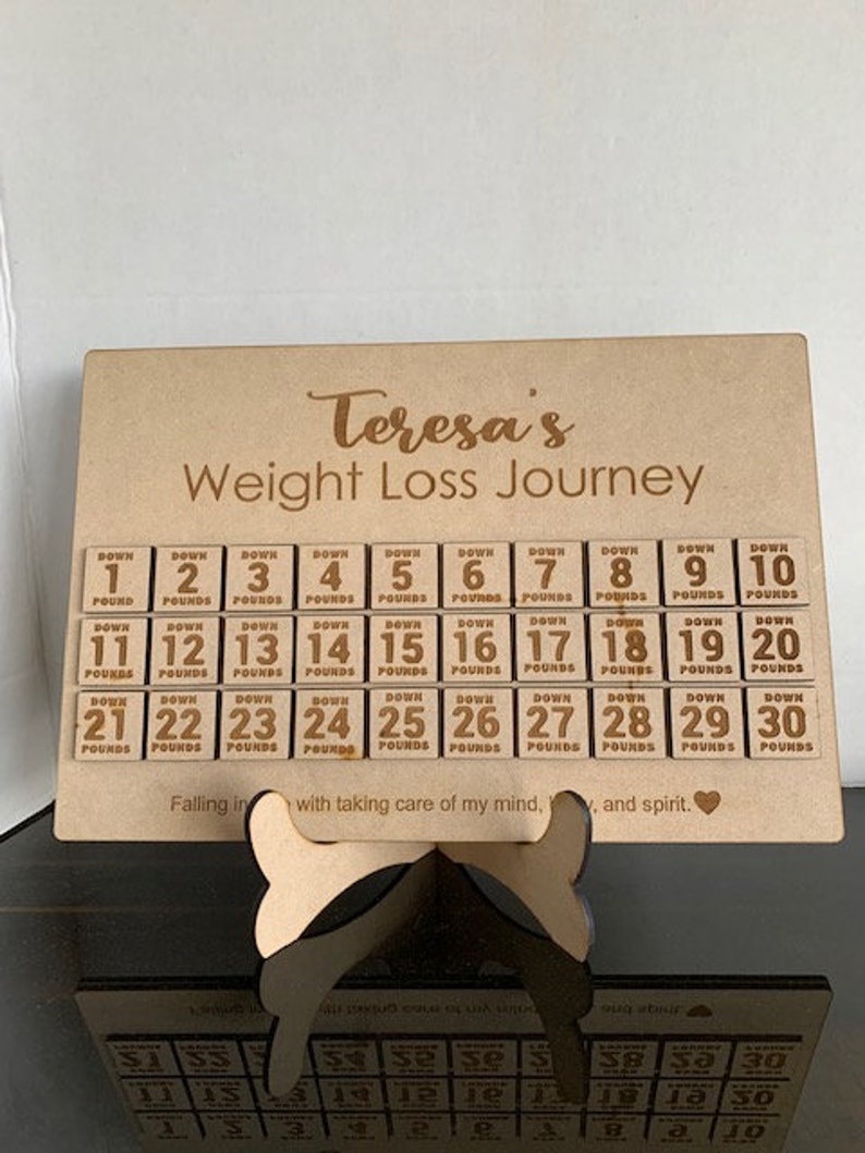 May include: A wooden board with the text "Teresa's Weight Loss Journey" and a grid of 30 small wooden blocks numbered 1 through 30. Each block has the word "Down" and "Pounds" printed on it. The board has the text "Falling in with taking care of my mind, and spirit."