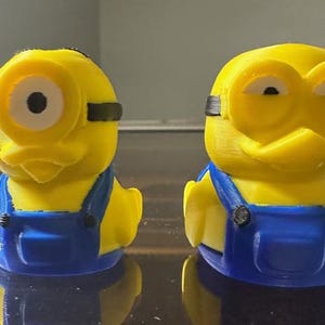 May include: Two yellow Minion-themed figurines. Each figure has blue overalls, black goggles, and a detailed face. The figures are standing upright, showcasing the design and color scheme. The background is blurred, focusing attention on the Minion figures.