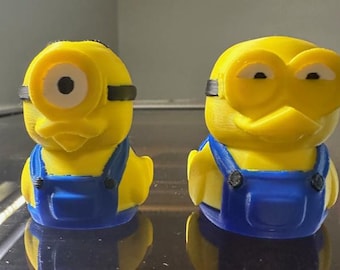 May include: Two yellow Minion-themed figurines. Each figure has blue overalls, black goggles, and a detailed face. The figures are standing upright, showcasing the design and color scheme. The background is blurred, focusing attention on the Minion figures.