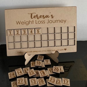 May include: A wooden weight loss tracker with the text "Teresa's Weight Loss Journey" and a grid of squares for tracking progress. The tracker comes with small wooden tiles with numbers and the word "Down" printed on them. The tiles are scattered around the tracker.