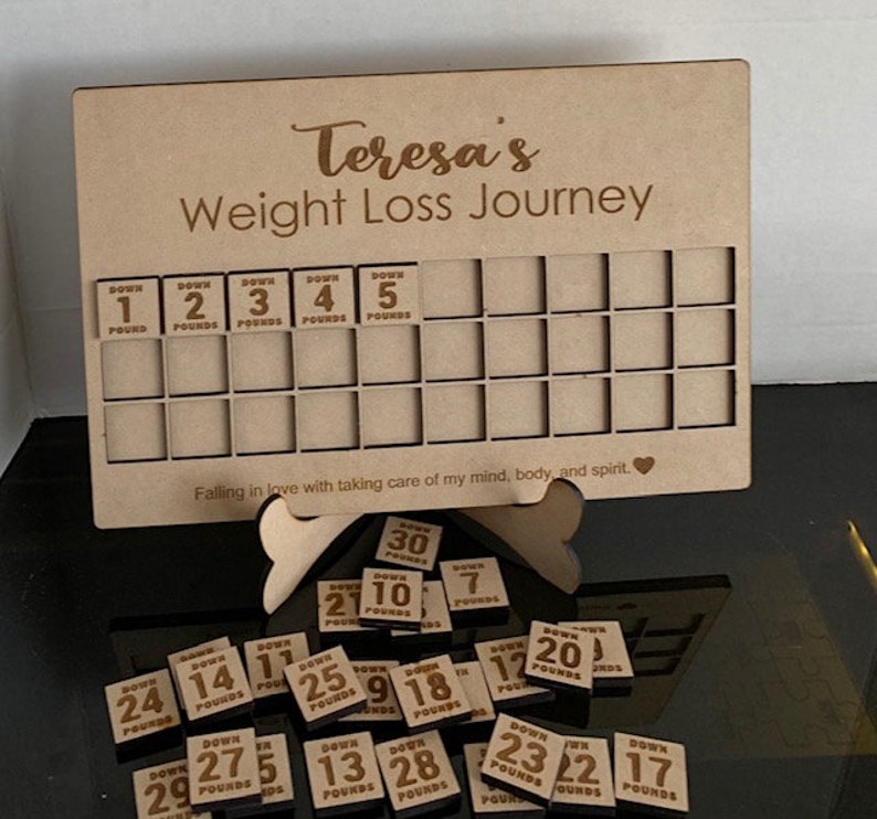 May include: A wooden weight loss tracker with the text "Teresa's Weight Loss Journey" and a grid of squares for tracking progress. The tracker comes with small wooden tiles with numbers and the word "Down" printed on them. The tiles are scattered around the tracker.