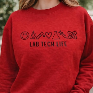 May include: A red sweatshirt with the text "LAB TECH LIFE" and various lab-related icons in black. The icons include a smiley face, microscope, test tube, heart, flask, syringe, and bandage. The sweatshirt is a solid red color.