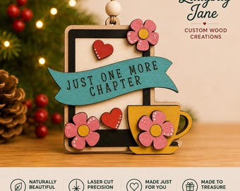 May include: A wooden hanging ornament featuring a rectangular frame with a teal banner printed with the text JUST ONE MORE CHAPTER. The design includes a yellow teacup, two red hearts, and two pink flowers with yellow centres, all suspended by a string.