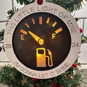 May include: A white wooden Christmas ornament shaped like a gas gauge with the text "This little light of mine, I'm gonna let it shine". The gauge is filled with a yellow light and has the letters "E" and "F" on either side.