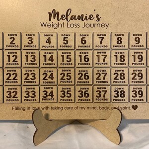 May include: A wooden weight loss tracker with a heart at the bottom. The tracker has 40 squares, each with the text "Down" and a number from 1 to 40, followed by the word "Pounds". The text "Melanie's Weight Loss Journey" is at the top of the tracker. The text "Falling in love with taking care of my mind, body, and spirit." is below the squares.