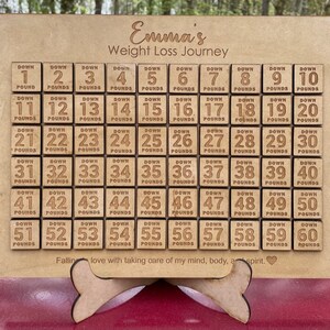 May include: A wooden weight loss tracker with a personalized name "Emma's Weight Loss Journey". The tracker has 60 squares, each with a number from 1 to 60 and the words "Down Pounds".