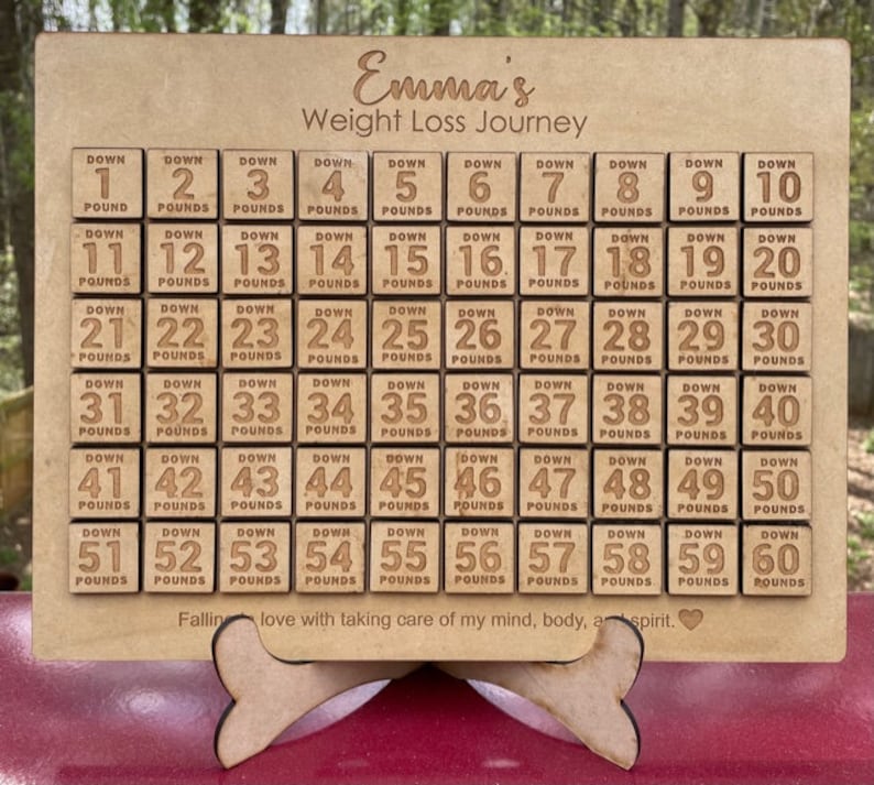 May include: A wooden weight loss tracker with a personalized name "Emma's Weight Loss Journey". The tracker has 60 squares, each with a number from 1 to 60 and the words "Down Pounds".
