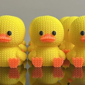May include: A group of yellow duck figurines with orange beaks and feet. The ducks have black eyes and a textured, knitted appearance. The figurines are arranged closely together, reflecting on a glossy surface.