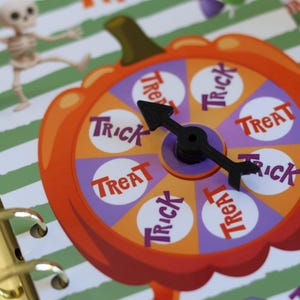 May include: A pumpkin-shaped spinner game featuring alternating orange and purple segments labelled with the words Trick and Treat. A black plastic arrow is attached to the centre. The game is set against a white and green striped background.