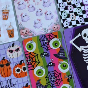 May include: A collection of rectangular cards featuring Halloween-themed illustrations including ghosts with bows, pumpkins with sunglasses, eyeballs, spider webs, spiders, witches' hats, and a skeleton inside a purple coffin shape.