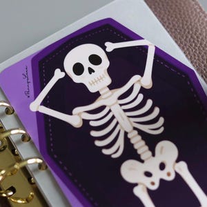May include: A rectangular sticker featuring a white skeleton lying inside a dark purple coffin. The skeleton has its arms raised behind its head. The sticker includes the text Penny Luna in a cursive script on the left side of the purple border.