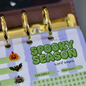 May include: A planner insert featuring a light purple and white striped background with the green text SPOOKY SEASON and Word Search. The page includes icons of a pumpkin, a bat, and a spider, along with pound symbols and a grid for word searching.