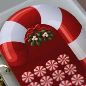 May include: A glossy planner divider featuring a large red and white striped candy cane graphic with a green mistletoe sprig tied with a red bow, positioned above a pattern of small red and white peppermint candies on a deep red background.