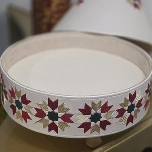 May include: A round, off-white decorative tray with a raised edge featuring an embroidered geometric star pattern in shades of deep red, dark green, and metallic gold. The interior base is smooth and off-white, and the tray stands on small feet.