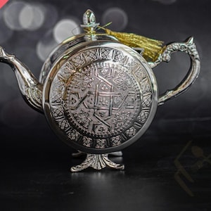 May include: A silver-toned metal teapot featuring an intricate circular embossed pattern on its side, a curved handle, a pointed spout, and a small decorative gold-colored tassel attached to the lid finial, standing on a base.