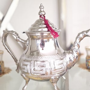 May include: A polished silver-colored teapot featuring a rounded body with an embossed Greek key pattern, a curved handle, a long spout, and a lid topped with a finial. A small, bright red decorative tassel is tied to the lid's handle.