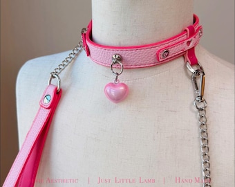 May include: A light pink textured choker collar featuring a central silver-toned ring with a dangling pearlescent pink heart charm. The collar includes silver-toned metal hardware, a matching pink strap, and silver chains. Text reads: THE AESTHETIC | JUST LITTLE LAMB | HAND MADE.