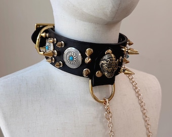 May include: A black leather choker collar featuring gold-toned metal spikes, silver-toned circular concho accents with central turquoise stones, and a gold-toned metal lion head ornament. A gold-toned metal D-ring attaches to a long metal chain.