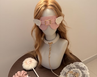 May include: A light pink blindfold with a gold buckle and white wing accents, worn on a mannequin head with blonde hair. A white faux fur collar with a gold chain leash, a white fluffy wand, and a pink ruffled scrunchie are also included.