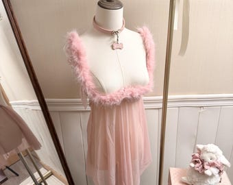 May include: A light pink sheer mesh babydoll dress featuring faux fur trim along the neckline and shoulder straps. The garment is displayed on a mannequin wearing a matching light pink collar with a bone-shaped pendant attached to a metal ring.