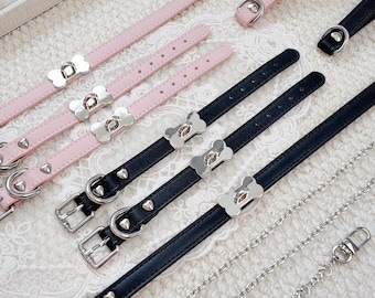 May include: A collection of thin, adjustable straps in pastel pink and black, each featuring silver-toned metal hardware, including bone-shaped accents, buckles, and D-rings. A silver-toned metal chain with small heart-shaped links is also included.