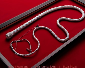 May include: A long, braided whip featuring a light beige and white pattern with a textured handle and a thin, flexible lash. The whip is displayed within a dark wooden frame. Text at the bottom reads: SOFTIE AESTHETIC | JUST LITTLE LAMB | HAND MADE.