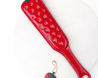 May include: A glossy red paddle featuring a quilted texture with small gold studs and a dark green leaf accent at the top. The handle includes a gold-toned metal eyelet at the base. A matching strawberry-shaped keychain with a gold chain is included.