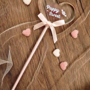 May include: A clear heart-shaped wand featuring the white text Puppy Sweet, accented with a light pink satin ribbon bow tied at the base of the heart. The wand is surrounded by small, fuzzy, heart-shaped embellishments in white and pink.