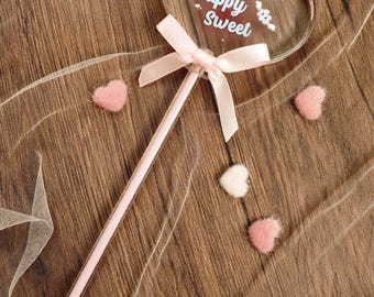 May include: A clear heart-shaped wand featuring the white text Puppy Sweet, accented with a light pink satin ribbon bow tied at the base of the heart. The wand is surrounded by small, fuzzy, heart-shaped embellishments in white and pink.