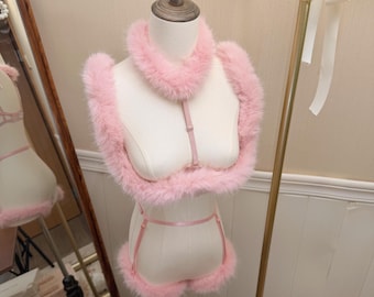 May include: A light pink faux fur lingerie set displayed on a mannequin. The set includes a collar, a cropped top with fur trim, and matching bottoms with fur-trimmed leg openings, all connected by thin, adjustable pink satin straps and metal sliders.