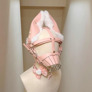May include: A light pink head harness featuring tall, pointed ears lined with white faux fur. The structure includes adjustable straps, silver-toned metal hardware, and a front-facing wire cage muzzle. A matching pink bow with a white heart sits below.