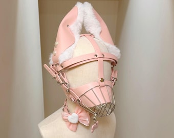 May include: A light pink head harness featuring tall, pointed ears lined with white faux fur. The structure includes adjustable straps, silver-toned metal hardware, and a front-facing wire cage muzzle. A matching pink bow with a white heart sits below.