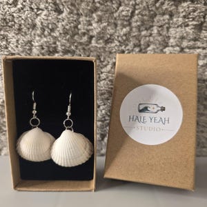 May include: A pair of white seashell earrings with silver-toned metal hooks, presented in a brown cardboard box with a black velvet insert. The box lid features a circular white sticker with a bottle graphic and the text HALE YEAH STUDIO.