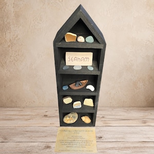 May include: A black wooden display shelf shaped like a house with five levels containing various sea glass pieces, small stones, shells, and a miniature brown boat. A rectangular sign reads SEAHAM. A separate card below reads COASTAL CURIOSITY HOUSE.