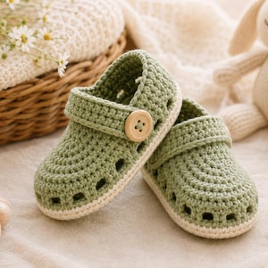 May include: A pair of sage green crochet baby shoes featuring a rounded toe, a side strap with a single light wood button, and a cream-colored sole. The shoes have a textured knit pattern with small cutouts along the sides for ventilation.